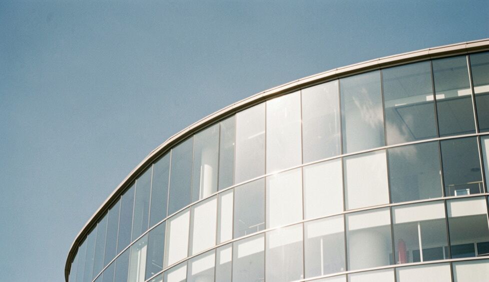 Fachada curva de vidrio reflectante bajo un cielo azul claro, con reflejos suaves sobre el metal blanco y líneas arquitectónicas limpias al fondo de la plaza.