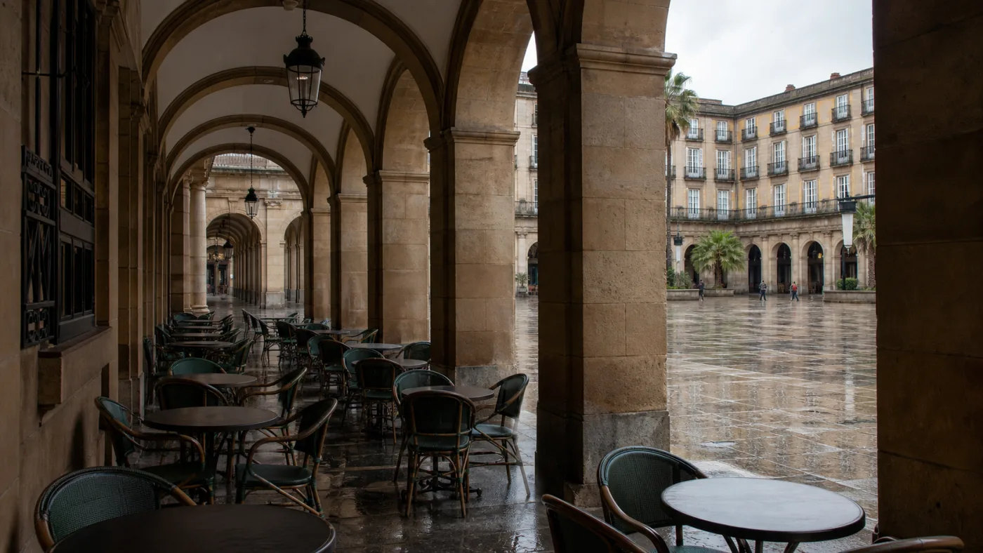 Arcadas de piedra de la Plaza Nueva después de la lluvia, con terrazas recogidas y el pavimento de la plaza reflejando el cielo.