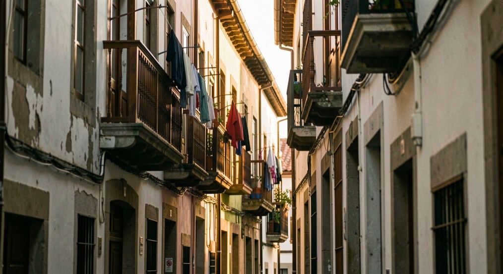 Calle ancha de barrio popular en Bilbao con fachadas grises y ocres, toldos gastados, peatones lejanos y una franja de sol entrando entre los edificios.